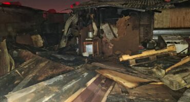 Damaged structure with burnt wooden beams and debris scattered on the ground, indicating a recent fire incident at night.