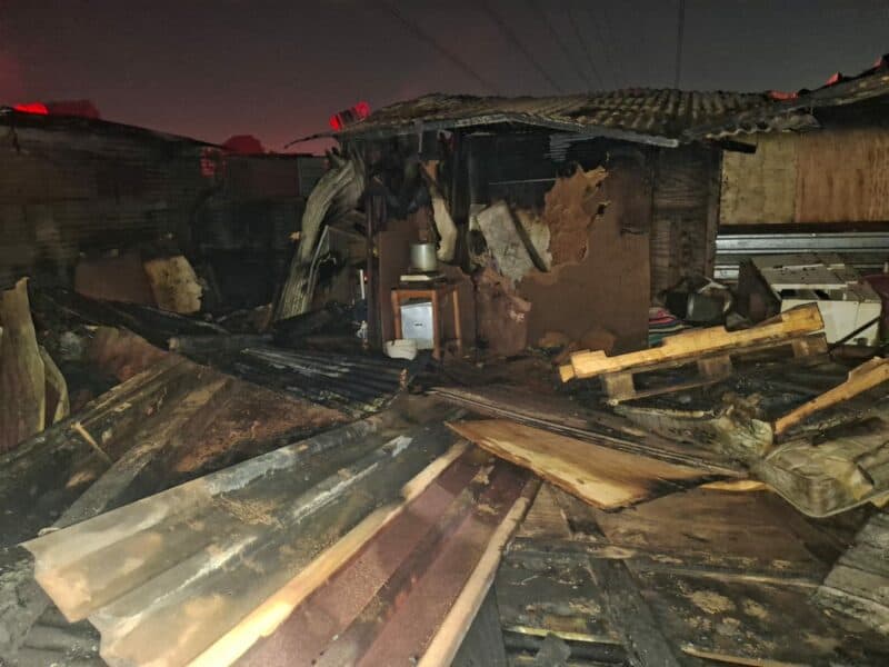 Damaged structure with burnt wooden beams and debris scattered on the ground, indicating a recent fire incident at night.