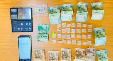 A collection of South African banknotes, coins, a digital scale, and small plastic bags arranged on a wooden surface.