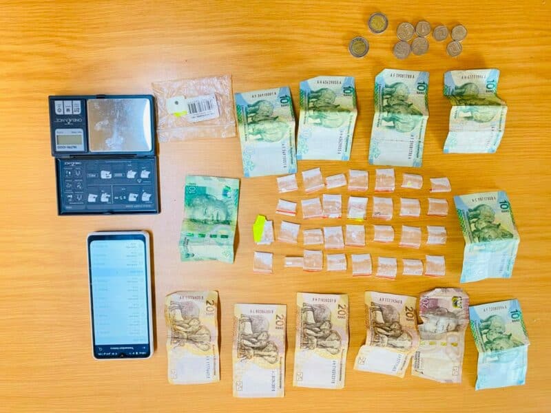 A collection of South African banknotes, coins, a digital scale, and small plastic bags arranged on a wooden surface.