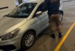 A security officer in a black jacket with 'EMPD' is inspecting a silver car parked in a garage with trees in the background.