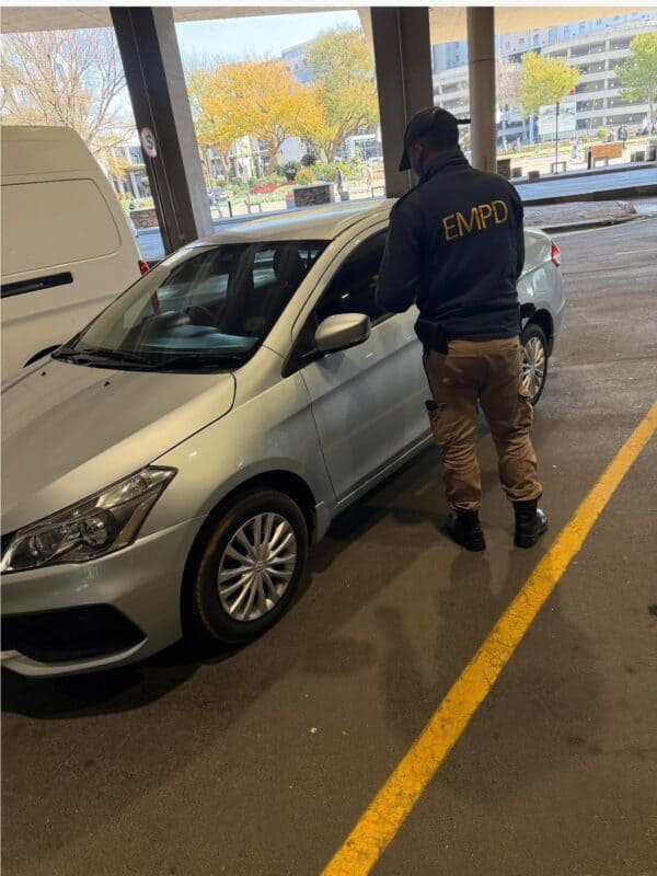 A security officer in a black jacket with 'EMPD' is inspecting a silver car parked in a garage with trees in the background.