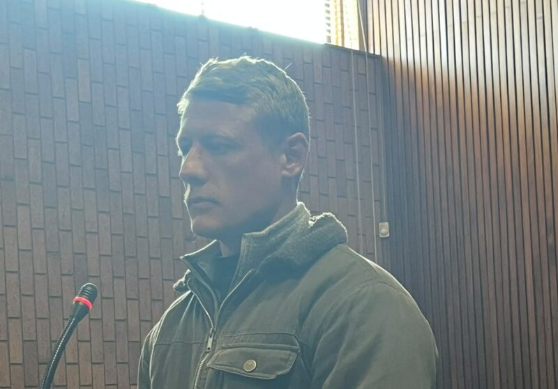 A man with short hair and a mustache stands in a courtroom, wearing a green jacket, with a microphone and wooden bench visible.