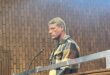 A man with short blonde hair wearing a brown and beige plaid jacket stands in a courtroom with a wood-paneled wall and a metal railing in front.