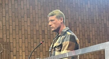 A man with short blonde hair wearing a brown and beige plaid jacket stands in a courtroom with a wood-paneled wall and a metal railing in front.