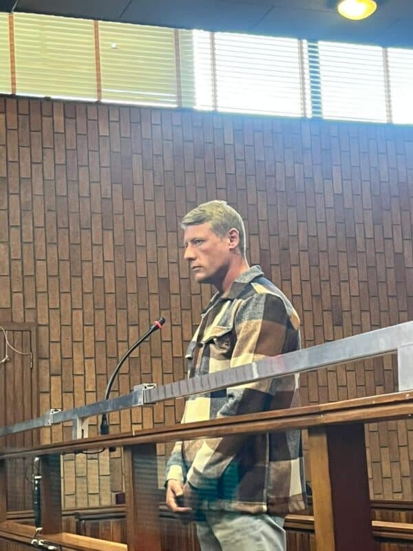 A man with short blonde hair wearing a brown and beige plaid jacket stands in a courtroom with a wood-paneled wall and a metal railing in front.