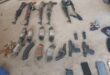 A collection of rifles, handguns, ammunition magazines, loose bullets, and two smartphones laid out on a concrete floor in a organized arrangement.