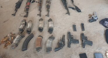 A collection of rifles, handguns, ammunition magazines, loose bullets, and two smartphones laid out on a concrete floor in a organized arrangement.