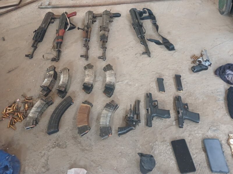 A collection of rifles, handguns, ammunition magazines, loose bullets, and two smartphones laid out on a concrete floor in a organized arrangement.