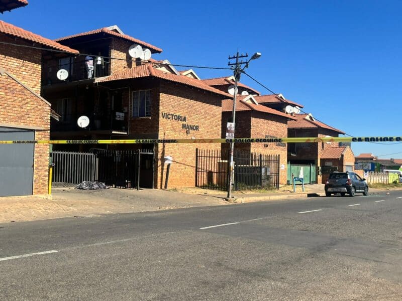 A brick apartment building named Victoria Manor cordoned off with yellow South African Police Service tape under a clear blue sky on a sunny day.