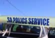 A strip of yellow police tape with the words SA POLICE SERVICE printed in bold blue letters, held in front of a white pickup truck against a blue sky.