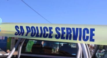 A strip of yellow police tape with the words SA POLICE SERVICE printed in bold blue letters, held in front of a white pickup truck against a blue sky.