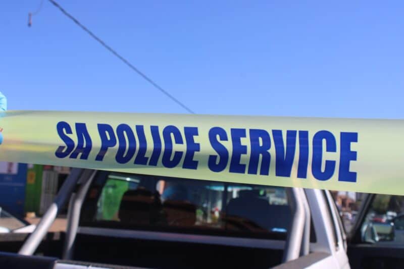 A strip of yellow police tape with the words SA POLICE SERVICE printed in bold blue letters, held in front of a white pickup truck against a blue sky.