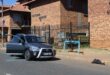 A grey Toyota parked on a street in front of Victoria Manor brick apartments with a broken window and glass scattered on the asphalt road surface.