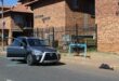 A grey Toyota parked on a street in front of Victoria Manor brick apartments with a broken window and glass scattered on the asphalt road surface.