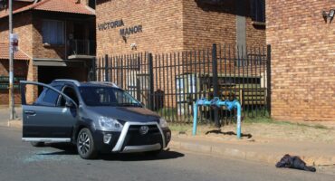 A grey Toyota parked on a street in front of Victoria Manor brick apartments with a broken window and glass scattered on the asphalt road surface.