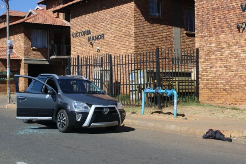 A grey Toyota parked on a street in front of Victoria Manor brick apartments with a broken window and glass scattered on the asphalt road surface.