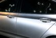 A silver sedan parked at night features multiple circular impact holes and spiderweb-like cracks across its dark, tinted side passenger windows.
