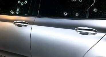 A silver sedan parked at night features multiple circular impact holes and spiderweb-like cracks across its dark, tinted side passenger windows.