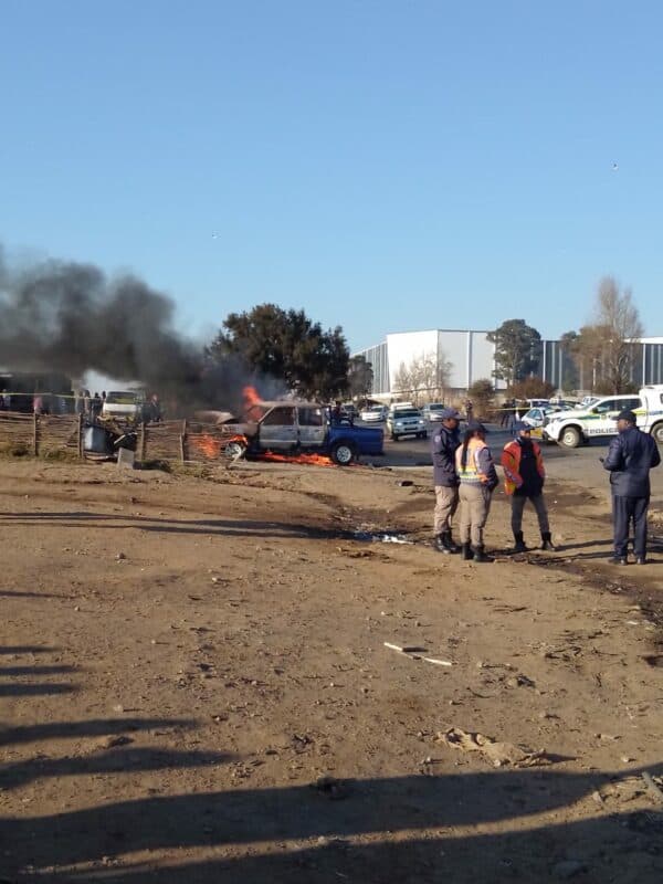 A blue pickup truck is engulfed in flames and smoke near a fence, while several police officers in uniform stand on a dirt road observing the scene.