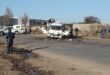A damaged white cash-in-transit truck sits on a road with police officers nearby, debris scattered on the ground, and a clear blue sky overhead.