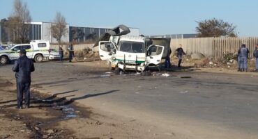 A damaged white cash-in-transit truck sits on a road with police officers nearby, debris scattered on the ground, and a clear blue sky overhead.