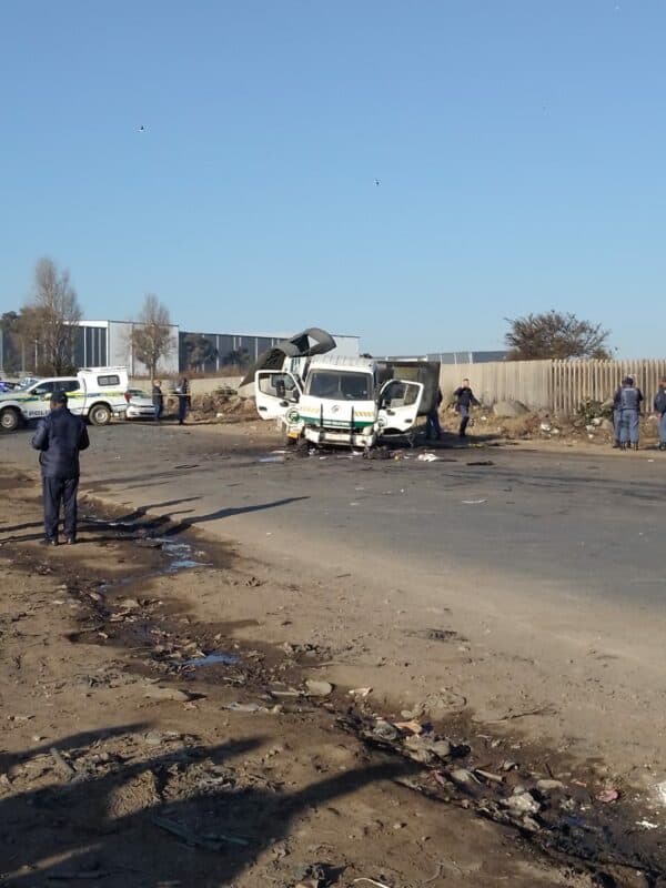 A damaged white cash-in-transit truck sits on a road with police officers nearby, debris scattered on the ground, and a clear blue sky overhead.
