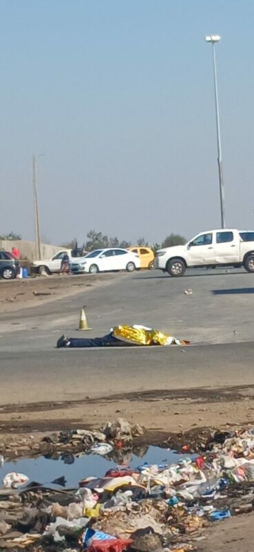 A person lying on an asphalt road covered by a reflective gold emergency blanket, with a traffic cone nearby and several cars parked in the background.