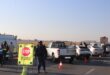 South African police officers conduct a roadside checkpoint on a paved road, with a stop sign, orange traffic cones, and several vehicles present.