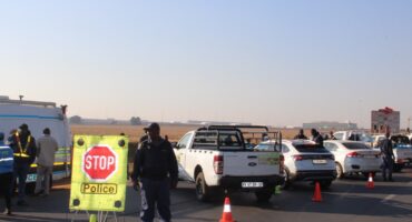 South African police officers conduct a roadside checkpoint on a paved road, with a stop sign, orange traffic cones, and several vehicles present.