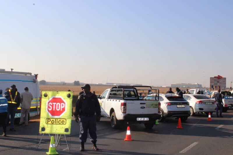 South African police officers conduct a roadside checkpoint on a paved road, with a stop sign, orange traffic cones, and several vehicles present.