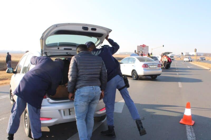 South African police officers conduct a roadside checkpoint on a paved road, with a stop sign, orange traffic cones, and several vehicles present.