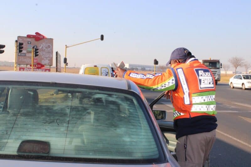 South African police officers conduct a roadside checkpoint on a paved road, with a stop sign, orange traffic cones, and several vehicles present.