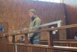 A man in a green jacket stands inside a wooden courtroom dock, facing forward with his hands clasped, while a woman sits in the background nearby.