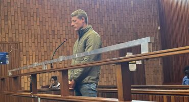 A man in a green jacket stands inside a wooden courtroom dock, facing forward with his hands clasped, while a woman sits in the background nearby.