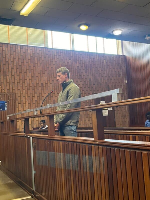 A man in a green jacket stands inside a wooden courtroom dock, facing forward with his hands clasped, while a woman sits in the background nearby.