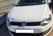 Front view of a white Volkswagen parked beside a highway on a sunny day, with a person standing nearby and long shadows on the gravel.