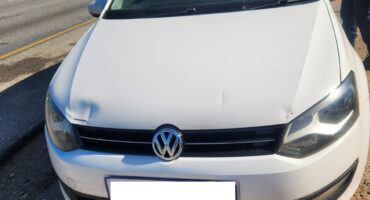 Front view of a white Volkswagen parked beside a highway on a sunny day, with a person standing nearby and long shadows on the gravel.