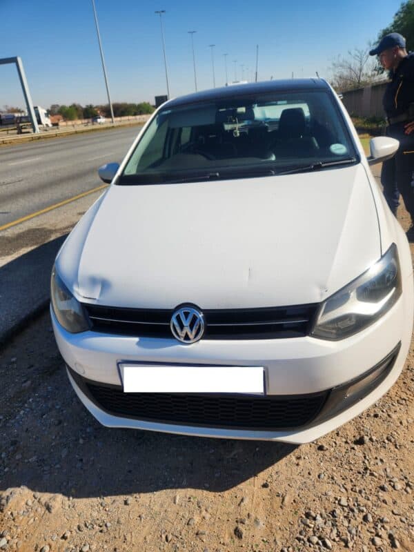 Front view of a white Volkswagen parked beside a highway on a sunny day, with a person standing nearby and long shadows on the gravel.