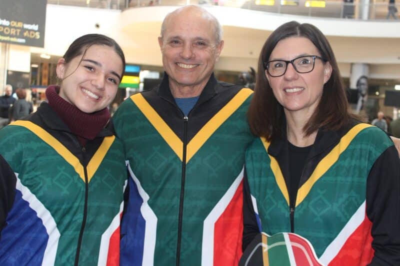 A young woman, an older man, and a middle-aged woman smile while wearing matching zip-up jackets featuring the South African flag color pattern.