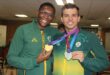 Two smiling men in green and yellow Team South Africa track jackets hold up gold and silver medals while posing together for a portrait indoors.