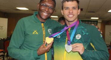 Two smiling men in green and yellow Team South Africa track jackets hold up gold and silver medals while posing together for a portrait indoors.