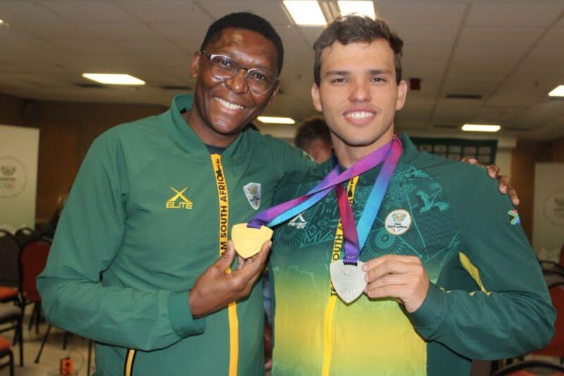 Two smiling men in green and yellow Team South Africa track jackets hold up gold and silver medals while posing together for a portrait indoors.