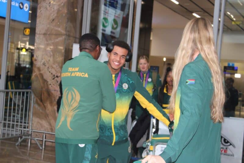 Athletes in green and yellow Team South Africa tracksuits stand in an airport terminal, with one smiling man wearing large black headphones.