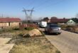 A semi-rural neighborhood with parked cars on a paved road. A utility tower stands in the background, near brick houses and dry grasslands under a clear sky.