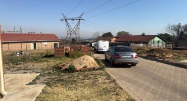 A semi-rural neighborhood with parked cars on a paved road. A utility tower stands in the background, near brick houses and dry grasslands under a clear sky.