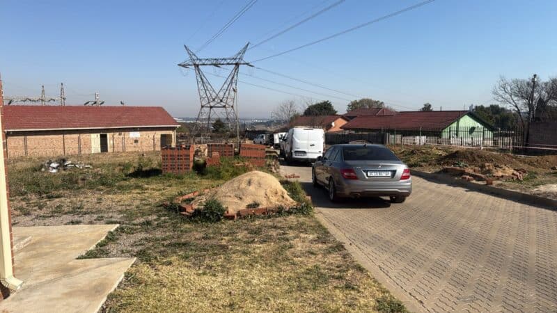 A semi-rural neighborhood with parked cars on a paved road. A utility tower stands in the background, near brick houses and dry grasslands under a clear sky.