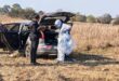 Two people beside an open SUV trunk in a dry field. One is in white protective gear, holding plastic bags. The mood is investigative and focused.