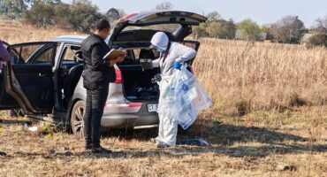 Two people beside an open SUV trunk in a dry field. One is in white protective gear, holding plastic bags. The mood is investigative and focused.