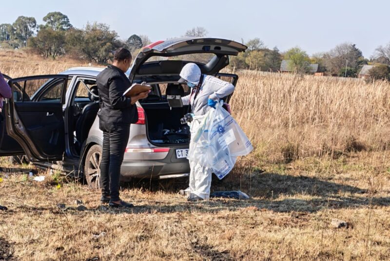 Two people beside an open SUV trunk in a dry field. One is in white protective gear, holding plastic bags. The mood is investigative and focused.